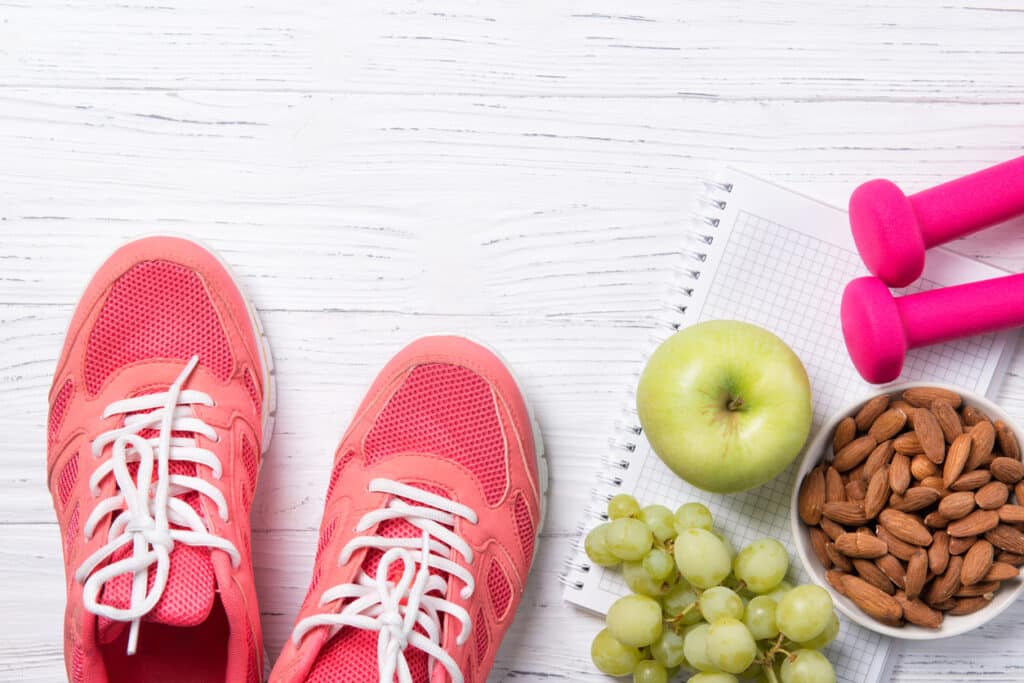 Fitness and healthy eating concept, pink sneakers and dumbbells with apple, grapes and almond nuts on notepad, wooden background