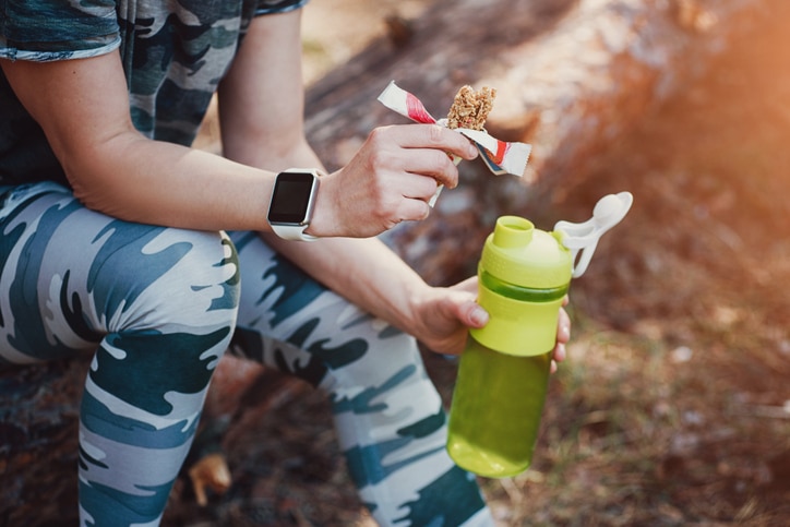 Sporty Woman Eating Protein Bar and drinking an amino acid cocktail. Woman in military-colored clothing in forest